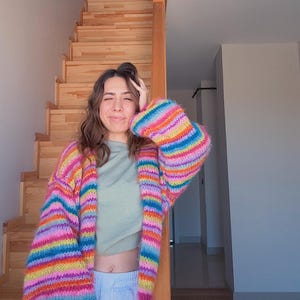 Könnte beinhalten: Ein farbenfroher, übergroßer Cardigan mit horizontalen Streifen in Rosa, Gelb, Blau und Grün. Der langärmlige Pullover wird über einem hellgrünen Oberteil und hellen Hosen getragen. Der Cardigan ist ein auffälliges Kleidungsstück.
