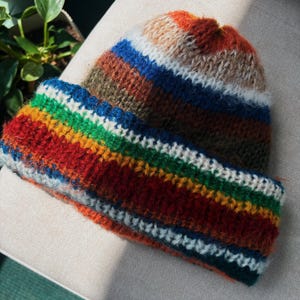 May include: A handmade, colorful knit hat with a folded brim. The hat features horizontal stripes in shades of red, orange, green, blue, and white. The hat is made of a soft, textured yarn, perfect for cold weather.