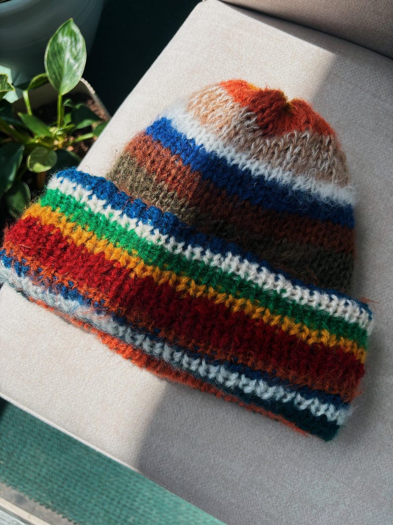 May include: A handmade, colorful knit hat with a folded brim. The hat features horizontal stripes in shades of red, orange, green, blue, and white. The hat is made of a soft, textured yarn, perfect for cold weather.