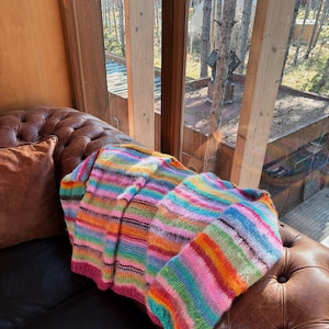 May include: A colorful striped knitted throw blanket with a variety of colors, including pink, blue, green, orange, and yellow, is draped over a brown leather couch.