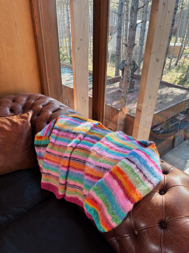 May include: A colorful striped knitted throw blanket with a variety of colors, including pink, blue, green, orange, and yellow, is draped over a brown leather couch.