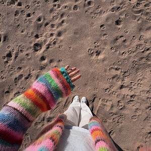 May include: A person wearing a colorful striped cardigan with a white shirt and white pants. The person is standing on a sandy beach.