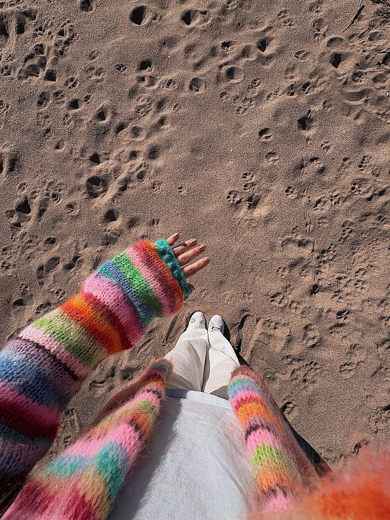 May include: A person wearing a colorful striped cardigan with a white shirt and white pants. The person is standing on a sandy beach.