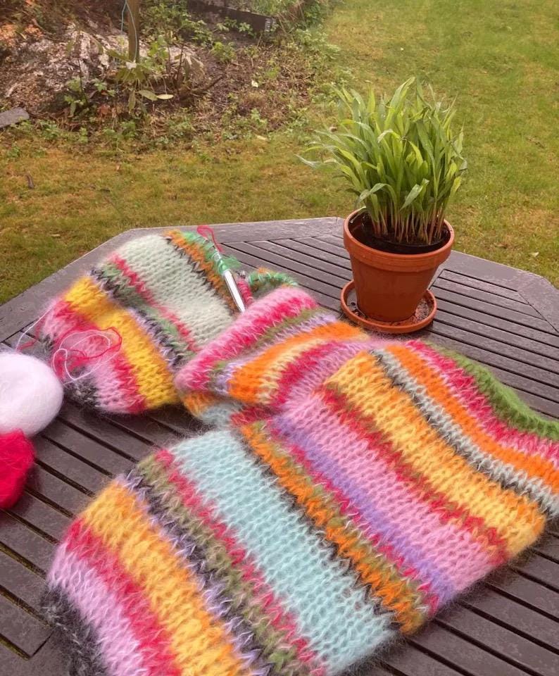 Puede incluir: Una bufanda de punto a rayas de colores con una variedad de colores que incluyen rosa, amarillo, verde y azul. La bufanda est&aacute; sobre una mesa de madera con una planta en maceta en el fondo.
