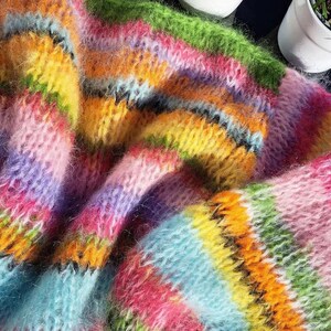 Puede incluir: Un primer plano de un su&eacute;ter de punto de rayas de colores con una textura peluda. El su&eacute;ter est&aacute; hecho de un hilo suave y esponjoso y presenta una variedad de colores brillantes, incluyendo rosa, azul, verde, amarillo y naranja.