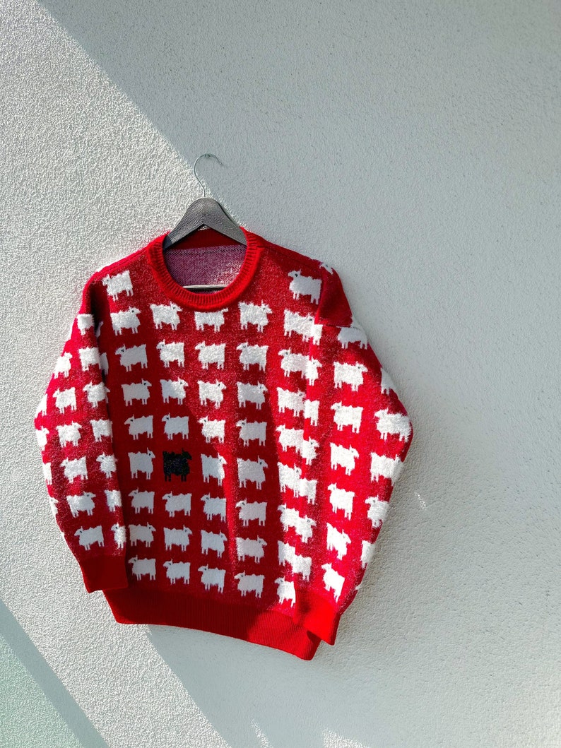Może przedstawiać: Czerwony sweter z wzorem białych owiec. Jedna czarna owca znajduje się w środku wzoru.