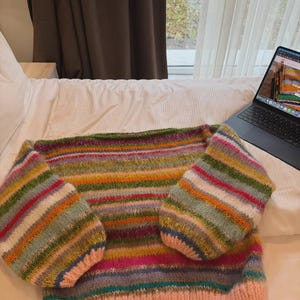 May include: A hand-knitted sweater with a striped pattern in various colors, including pink, green, orange, and blue. The sweater has a pink ribbed hem and cuffs, and is displayed on a white bed. The sleeves are wide and the body is a straight cut.