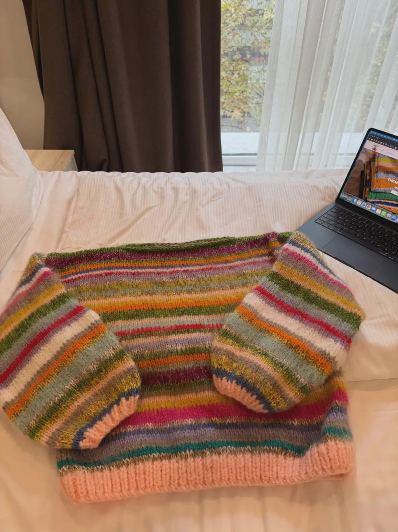May include: A hand-knitted sweater with a striped pattern in various colors, including pink, green, orange, and blue. The sweater has a pink ribbed hem and cuffs, and is displayed on a white bed. The sleeves are wide and the body is a straight cut.