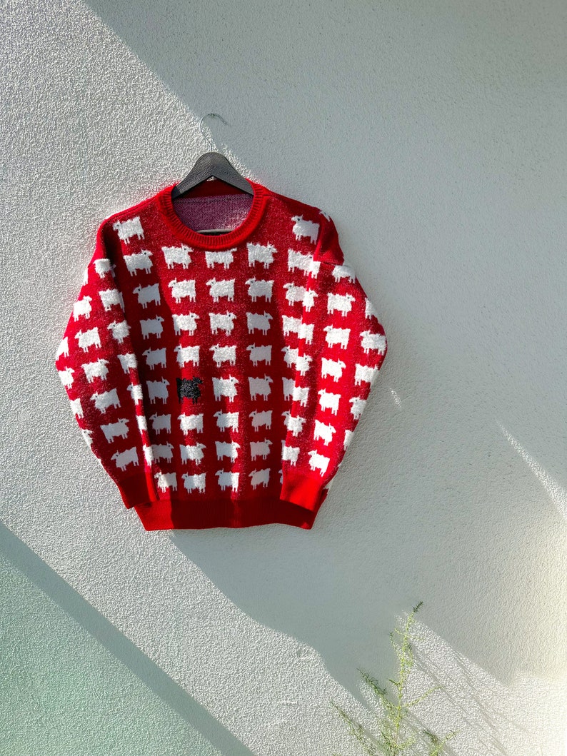 Może przedstawiać: Czerwony sweter z wzorem białych owiec. Jedna owca jest czarna.