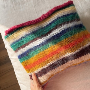 May include: A hand-knitted cowl with a vibrant striped pattern in shades of green, blue, yellow, orange, and brown. The top edge is finished with a bright pink border. The cowl is made of a soft, textured yarn.