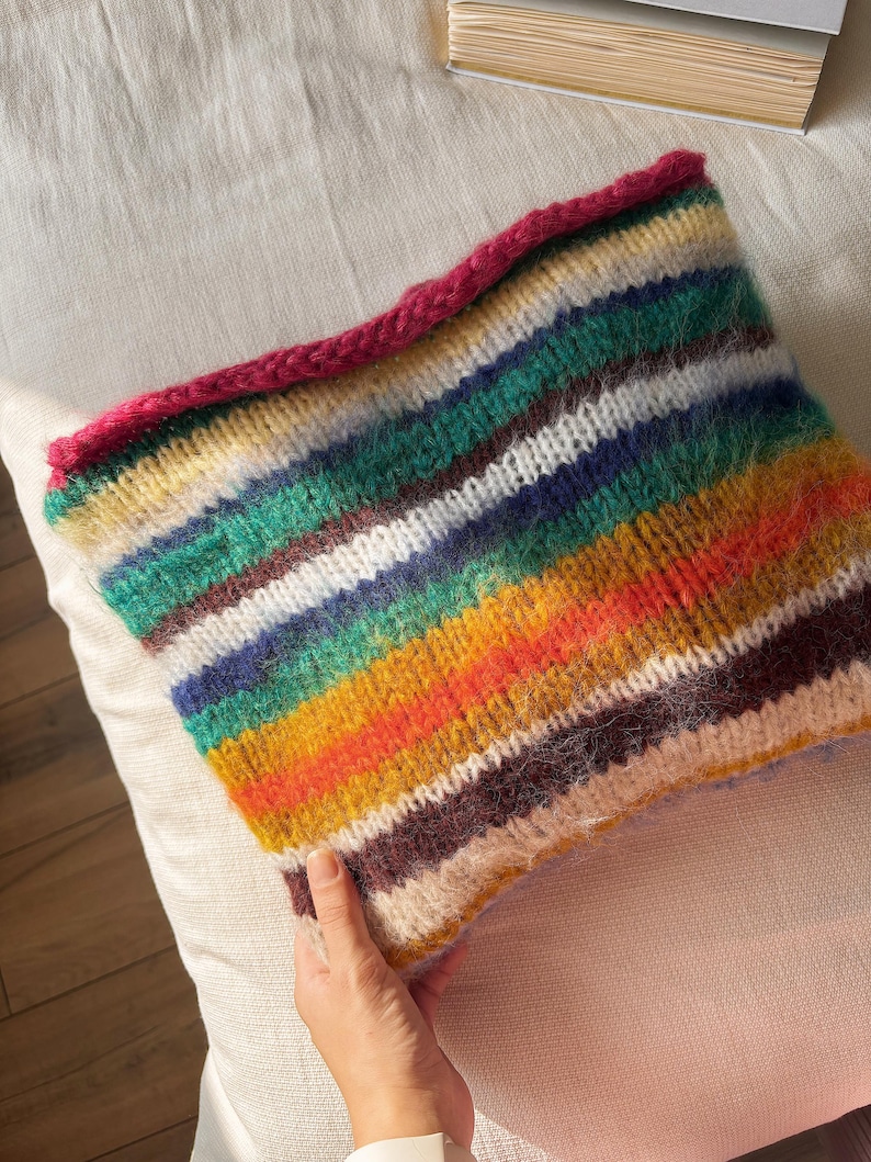 May include: A hand-knitted cowl with a vibrant striped pattern in shades of green, blue, yellow, orange, and brown. The top edge is finished with a bright pink border. The cowl is made of a soft, textured yarn.