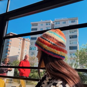 May include: A colorful, striped knit beanie in shades of red, orange, yellow, green, blue, and pink. The hat is worn by a person with long brown hair, paired with a black and white crochet top. The background shows a sunny day with buildings and people.