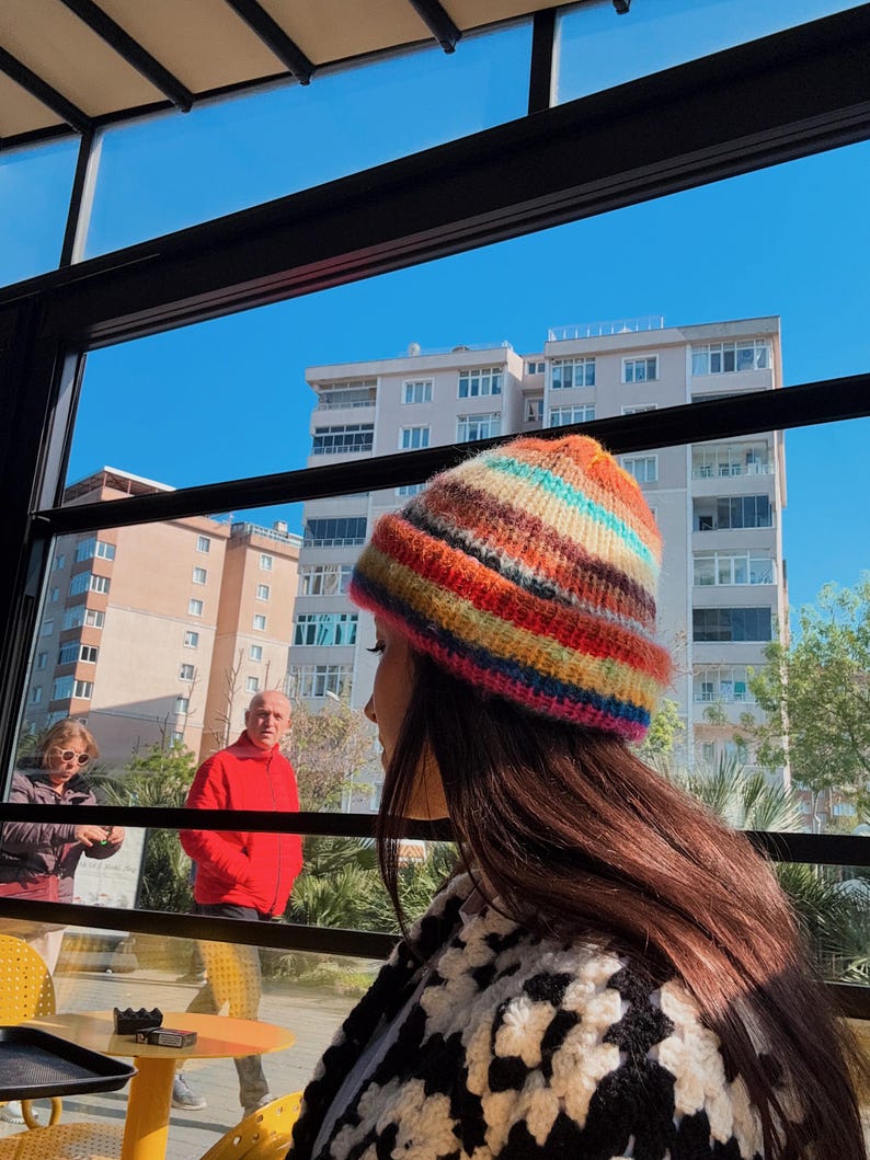 May include: A colorful, striped knit beanie in shades of red, orange, yellow, green, blue, and pink. The hat is worn by a person with long brown hair, paired with a black and white crochet top. The background shows a sunny day with buildings and people.