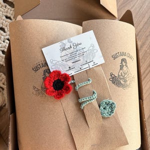 May include: A craft box with a red crocheted flower, a green crocheted spiral, and a green crocheted circle. The box contains rolled paper with the text "SUSTAIN CRAFT" and a thank you card with the text "Thank You".