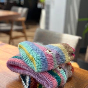 May include: A stack of colorful, hand-knitted beanies. Each beanie features a striped pattern in shades of pink, blue, yellow, and green. The beanies are made of a soft, textured yarn and are stacked on a wooden surface.