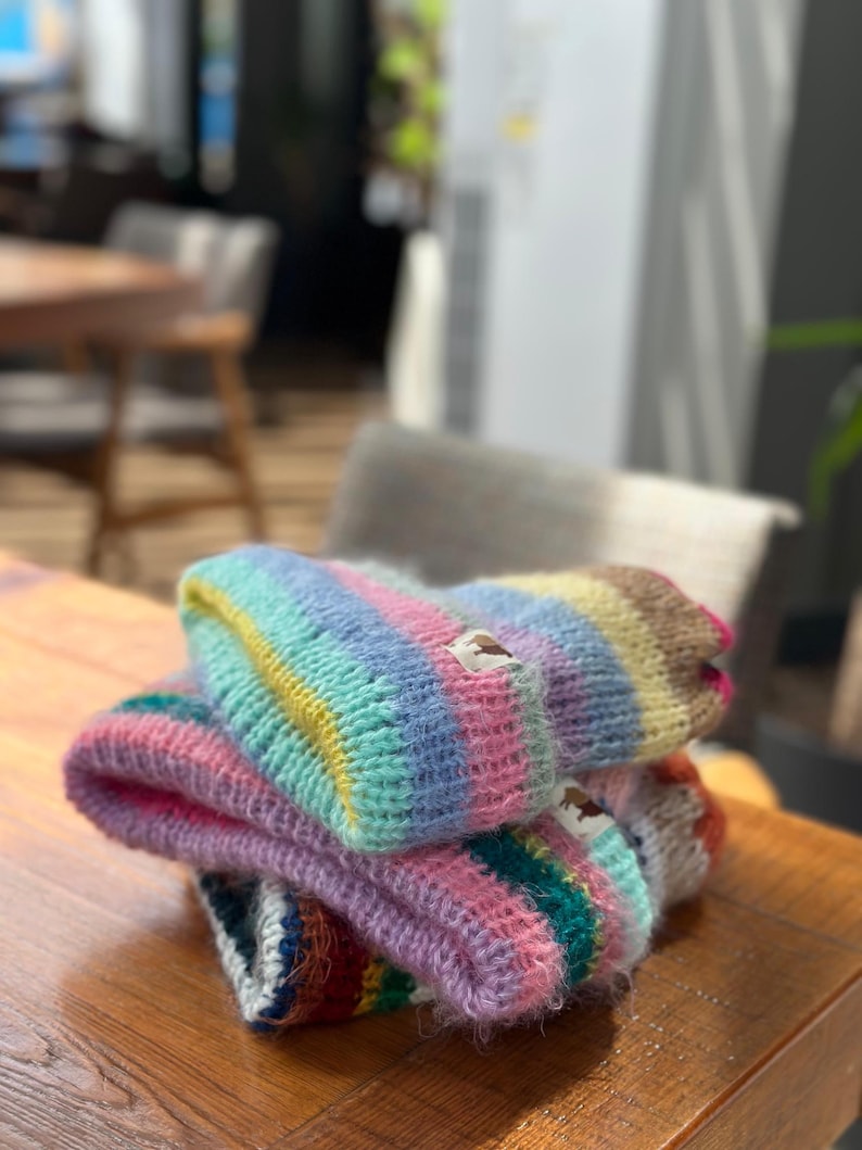 May include: A stack of colorful, hand-knitted beanies. Each beanie features a striped pattern in shades of pink, blue, yellow, and green. The beanies are made of a soft, textured yarn and are stacked on a wooden surface.
