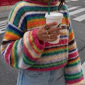 May include: A colorful, striped sweater with a rainbow of hues, including pink, green, yellow, and blue. The sweater has long sleeves and a round neckline. The person is wearing light blue jeans and holding a white coffee cup with a green straw.