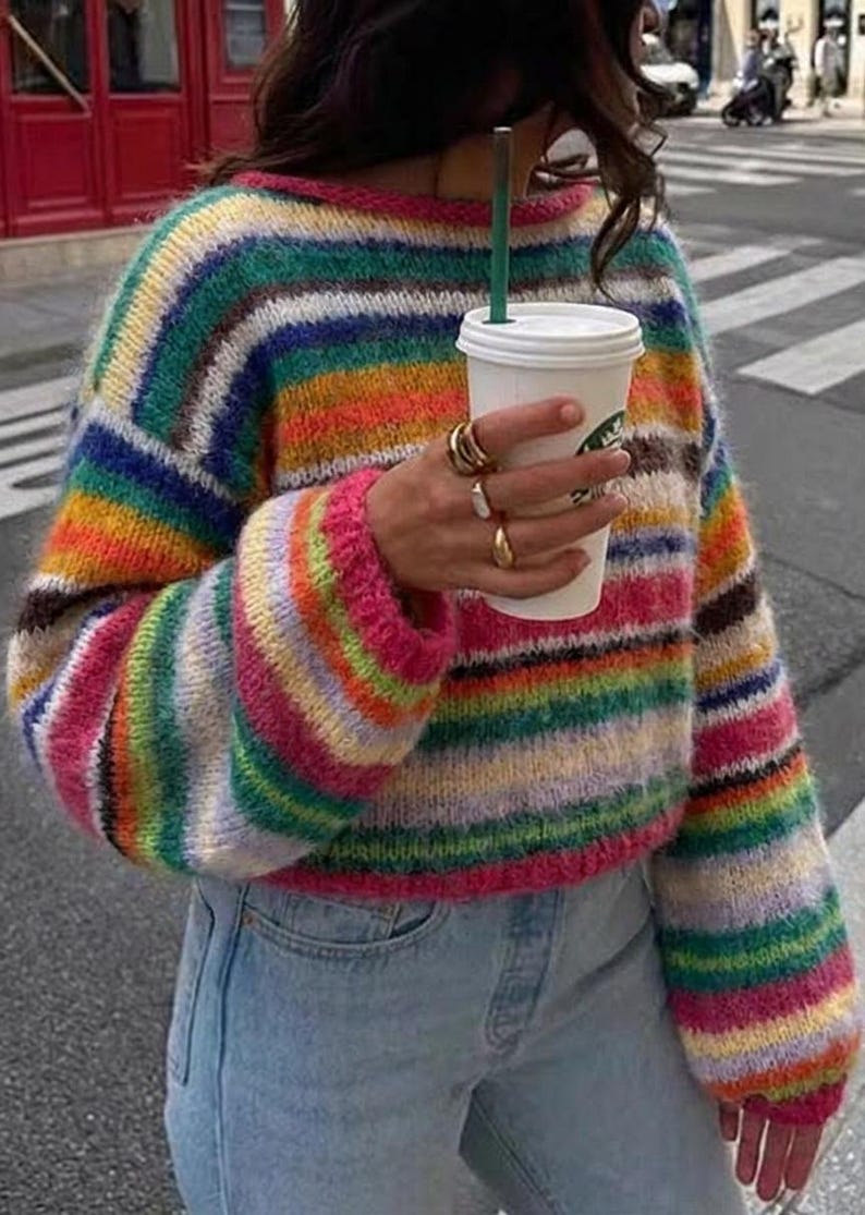 May include: A colorful, striped sweater with a rainbow of hues, including pink, green, yellow, and blue. The sweater has long sleeves and a round neckline. The person is wearing light blue jeans and holding a white coffee cup with a green straw.
