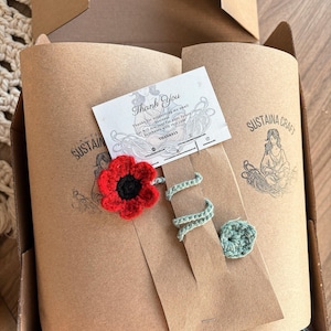 Może przedstawiać: Brązowe pudełko kartonowe z kartką z podziękowaniem w środku. Kartka ma na sobie czerwoną kwiatkę wykonaną na szydełku. Pudełko jest związane zieloną wstążką wykonaną na szydełku. Tekst na kartce brzmi: "Dziękuję" i "Dziękuję za wsparcie mojej małej firmy. Otrzymaj 15% zniżki na swoje przyszłe zakupy z tym kodem THANKS15".