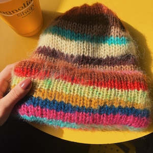 May include: A colorful, hand-knitted beanie hat with horizontal stripes in shades of red, orange, yellow, green, blue, and pink. The hat is made of a soft, textured yarn and has a warm, cozy appearance. The hat is sitting on a yellow surface.