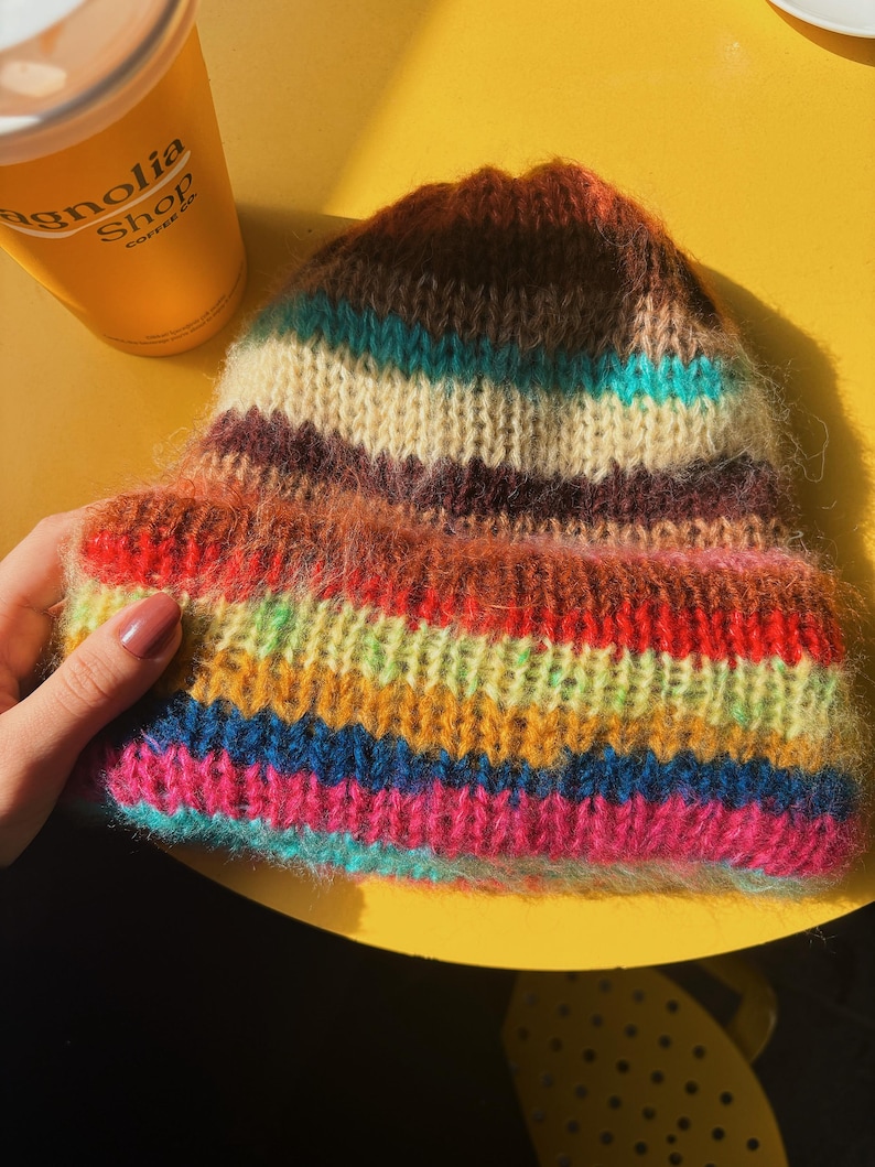 May include: A colorful, hand-knitted beanie hat with horizontal stripes in shades of red, orange, yellow, green, blue, and pink. The hat is made of a soft, textured yarn and has a warm, cozy appearance. The hat is sitting on a yellow surface.