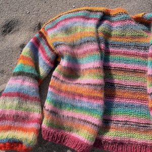 May include: A colorful striped knitted cardigan with a pink trim. The cardigan is made of a soft, fuzzy yarn and has a relaxed fit.