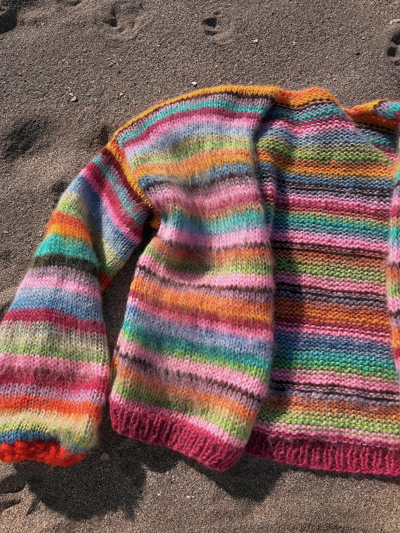 May include: A colorful striped knitted cardigan with a pink trim. The cardigan is made of a soft, fuzzy yarn and has a relaxed fit.