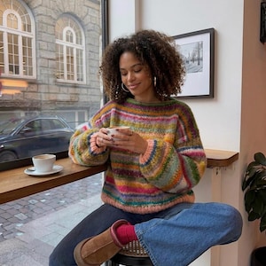 May include: A woman wearing a colorful striped sweater with a round neck and long sleeves, paired with blue jeans and brown boots. She is holding a white cup of coffee, seated near a window with a view of a city street.