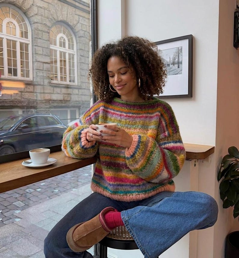 May include: A woman wearing a colorful striped sweater with a round neck and long sleeves, paired with blue jeans and brown boots. She is holding a white cup of coffee, seated near a window with a view of a city street.