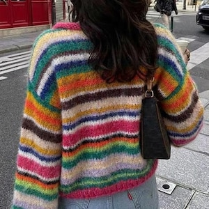 May include: A colorful, striped sweater in shades of pink, orange, green, blue, and brown. The sweater has long sleeves and a crew neck. The person is wearing light blue jeans and a black purse. The sweater is a cozy, casual style.