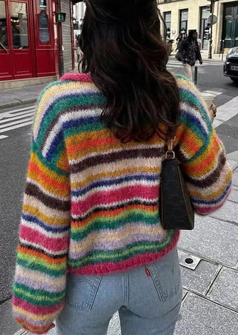 May include: A colorful, striped sweater in shades of pink, orange, green, blue, and brown. The sweater has long sleeves and a crew neck. The person is wearing light blue jeans and a black purse. The sweater is a cozy, casual style.