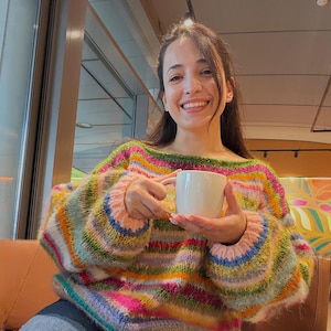 May include: A colorful, striped sweater with shades of pink, green, blue, and yellow. The sweater has a wide neckline and long sleeves. A white mug is held in the hands.