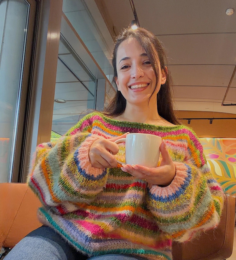 May include: A colorful, striped sweater with shades of pink, green, blue, and yellow. The sweater has a wide neckline and long sleeves. A white mug is held in the hands.