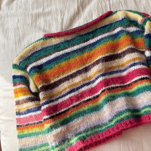May include: A colorful, striped sweater with a crew neck and long sleeves. The sweater features horizontal stripes in various colors, including pink, green, yellow, and blue. The neckline and cuffs are trimmed with a bright pink yarn.
