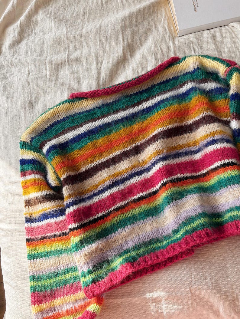 May include: A colorful, striped sweater with a crew neck and long sleeves. The sweater features horizontal stripes in various colors, including pink, green, yellow, and blue. The neckline and cuffs are trimmed with a bright pink yarn.