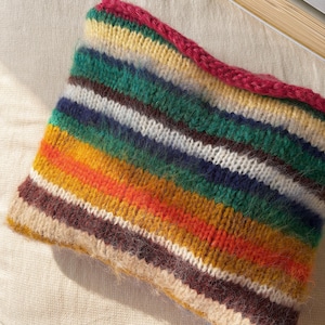 May include: A colorful, knitted neck warmer with horizontal stripes in shades of green, yellow, orange, brown, white, and blue. The top edge is a solid magenta color. The item is made of a soft, textured yarn.
