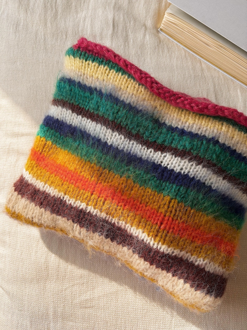 May include: A colorful, knitted neck warmer with horizontal stripes in shades of green, yellow, orange, brown, white, and blue. The top edge is a solid magenta color. The item is made of a soft, textured yarn.