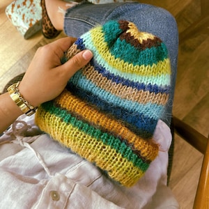 May include: A colorful, hand-knitted beanie hat with horizontal stripes of yellow, green, blue, and brown. The hat is held in a person's hand, showcasing the textured yarn and vibrant color palette. A gold watch is visible on the wrist.