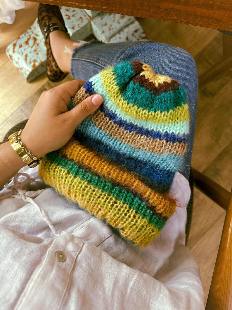 May include: A colorful, hand-knitted beanie hat with horizontal stripes of yellow, green, blue, and brown. The hat is held in a person's hand, showcasing the textured yarn and vibrant color palette. A gold watch is visible on the wrist.