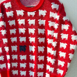 Może przedstawiać: Czerwony, puszysty sweter z wzorem białych owiec. Jedna czarna owca znajduje się w środku swetra.