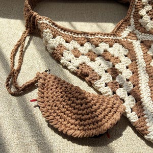 Puede incluir: Bolso y monedero de ganchillo hechos a mano en tonos marr&oacute;n y crema. El bolso presenta un dise&ntilde;o de cuadrados de la abuela y una correa larga. El monedero tiene forma de semic&iacute;rculo con cierre de cremallera y una etiqueta roja.