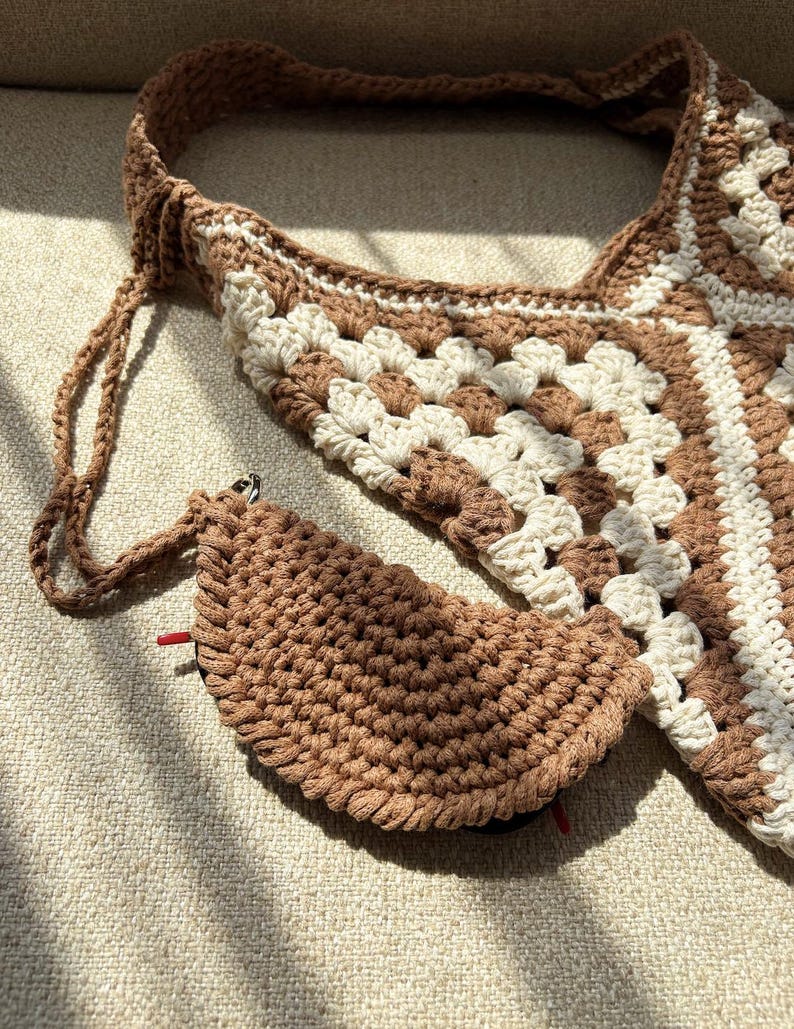 Puede incluir: Bolso y monedero de ganchillo hechos a mano en tonos marr&oacute;n y crema. El bolso presenta un dise&ntilde;o de cuadrados de la abuela y una correa larga. El monedero tiene forma de semic&iacute;rculo con cierre de cremallera y una etiqueta roja.