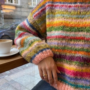 May include: A colorful, striped sweater with a round neckline and long sleeves. The sweater features a vibrant mix of colors including pink, green, yellow, and blue. The cuffs and neckline are a solid pink color. The sweater is made of a thick, textured yarn.