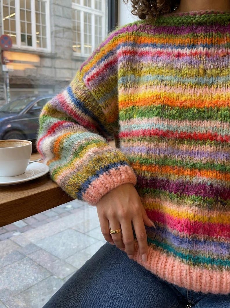 May include: A colorful, striped sweater with a round neckline and long sleeves. The sweater features a vibrant mix of colors including pink, green, yellow, and blue. The cuffs and neckline are a solid pink color. The sweater is made of a thick, textured yarn.