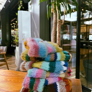 May include: A stack of colorful, hand-knitted mittens. The mittens are made of soft, fuzzy yarn in a variety of colors, including pink, blue, yellow, green, and orange. The mittens are stacked on a wooden table.