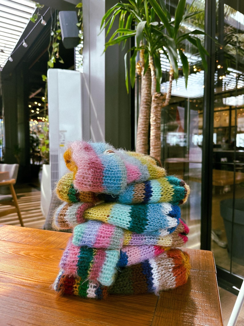 May include: A stack of colorful, hand-knitted mittens. The mittens are made of soft, fuzzy yarn in a variety of colors, including pink, blue, yellow, green, and orange. The mittens are stacked on a wooden table.