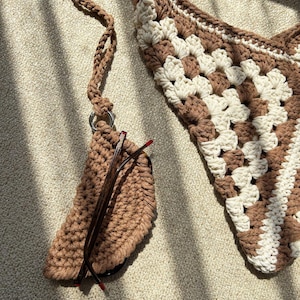 Puede incluir: Un bolso de hombro tejido a ganchillo marr&oacute;n y crema con un estuche para gafas a juego. El bolso presenta un dise&ntilde;o de granny square. El estuche para gafas est&aacute; sujeto al bolso con una correa de ganchillo marr&oacute;n y un anillo met&aacute;lico. El estuche para gafas contiene unas gafas.