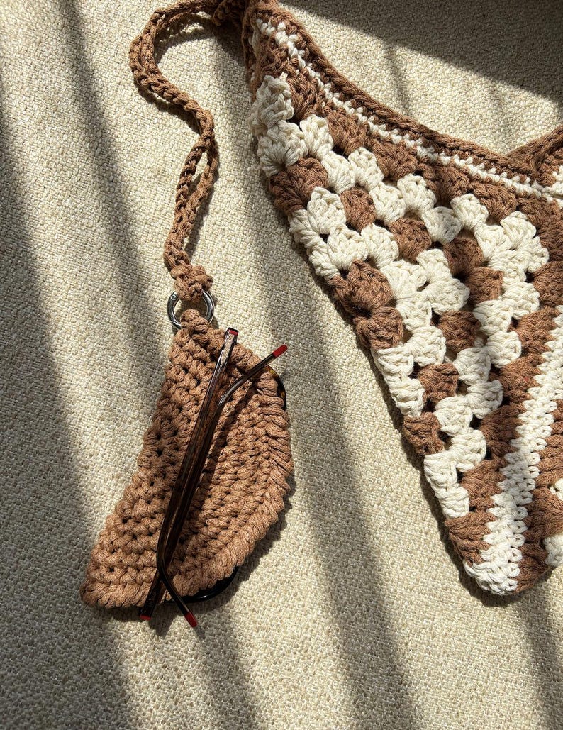 Puede incluir: Un bolso de hombro tejido a ganchillo marr&oacute;n y crema con un estuche para gafas a juego. El bolso presenta un dise&ntilde;o de granny square. El estuche para gafas est&aacute; sujeto al bolso con una correa de ganchillo marr&oacute;n y un anillo met&aacute;lico. El estuche para gafas contiene unas gafas.