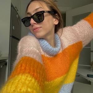 以下が含まれることがあります： 黒いサングラスとマルチカラーのストライプセーターを着た女性。セーターはピンク、オレンジ、イエロー、ブルーの横縞模様です。