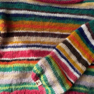 May include: A colorful, striped sweater with a matching sleeve. The sweater features horizontal bands of various colors, including pink, green, orange, and brown. The neckline and sleeve cuff are trimmed with a bright pink. The sweater is made of a soft, textured yarn.